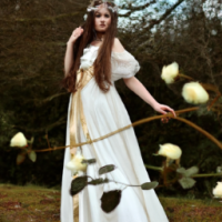 nature fairy girl with brown hair wearing a long flowy white dress with a golden belt and a crown o her haed decorated by white roses