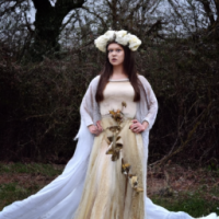 nature fairy girl with brown hair wearing a long flowy white dress with a golden belt and a crown on her head decorated by white roses